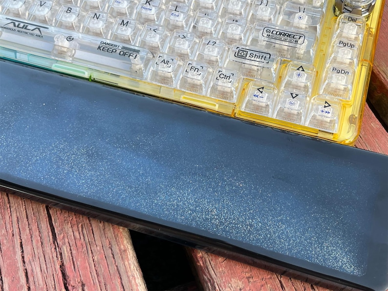 K&ouml;nnte beinhalten: Eine gelbe und transparente Tastatur aus Kunststoff mit schwarzen Beschriftungen. Die Tastatur hat ein schwarzes und silbernes Geh&auml;use mit einem Warnhinweis, der "DANGER! KEEP OFF!" lautet. Die Tastatur liegt auf einer Holzoberfl&auml;che.
