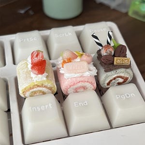 以下が含まれることがあります： 白いキーボードの上に置かれた3つのミニチュアケーキロール型キーキャップ。左はイチゴが乗った黄色いケーキ、中央はロータスビスケットが乗ったピンクのケーキ、右はコーヒー豆と白黒のストライプのストローが乗った茶色のケーキです。