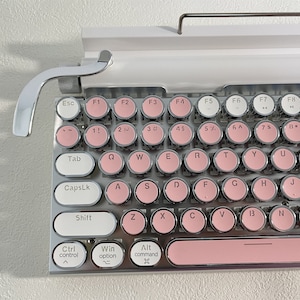 Pode incluir: Um teclado de estilo retro com moldura branca e teclas prateadas. As teclas redondas são de cor rosa claro com letras brancas. O teclado tem uma alavanca cromada no lado esquerdo.