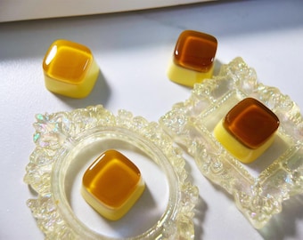 May include: Four small, square, yellow and brown resin keycaps with a glossy finish. The keycaps are arranged on a white surface with a decorative, iridescent frame.