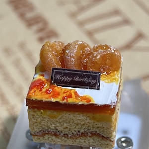 May include: A miniature square cake with layers of sponge, cream, and orange glaze, topped with three candied fruits. A small rectangular sign reads "Happy Birthday". The cake sits on a clear plastic stand.