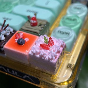 Può includere: Primo piano di un vassoio pieno di dessert in miniatura. Il vassoio contiene piccole torte quadrate con vari condimenti, tra cui fragole, mirtilli e panna montata. Altri articoli includono caramelle verdi e bianche. Il vassoio è in un contenitore trasparente color oro.