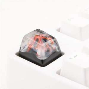 Può includere: Un keycap nero e bianco con un design a spirale rosso e bianco. Il keycap è realizzato in resina e ha una finitura lucida.
