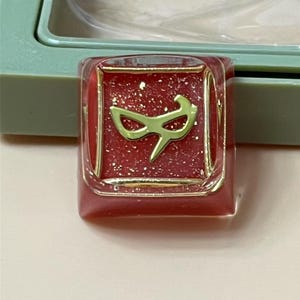 Op de afbeelding: Een vierkante, rode hars keycap met gouden glitter en een goudkleurig maskerontwerp. De keycap is gezet in een goudkleurig frame. De achtergrond is een lichtgroene plastic behuizing.