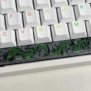 May include: A black and green keyboard keycap with a dragon design. The keycap is made of resin and has a textured surface.