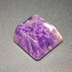May include: A purple amethyst crystal shaped like a keycap. The crystal has a smooth, polished surface and a cloudy, translucent appearance.
