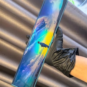 Op de afbeelding: Een rechthoekige smartphone met turquoise randen toont een levendige landschapsafbeelding met een regenboog en een figuur. De telefoon wordt vastgehouden door een gehandschoende hand, die het scherm benadrukt.