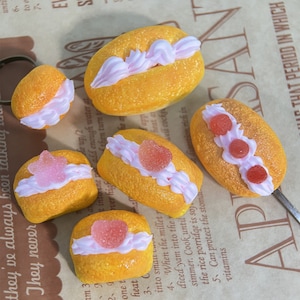 Può includere: Vista dall'alto di sei piccole torte gialle di forma ovale. Ogni torta è decorata con glassa bianca e una caramella rosa a forma di cuore o rotonda. Le torte sono disposte su una superficie marrone a motivi.