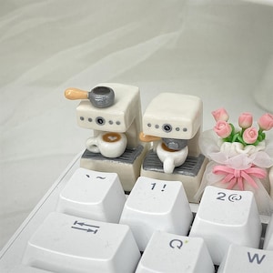 Puede incluir: Dos máquinas de espresso en miniatura blancas con un mango marrón y una taza de café con un diseño de corazón. Las máquinas están sobre un teclado blanco con teclas etiquetadas con "~", "!", "2@", "←", "Q" y "W".