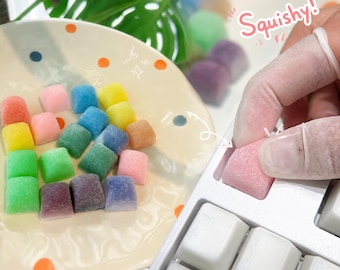 May include: A collection of small, colorful, textured square keycaps in shades of pink, yellow, blue, green, and purple. Some keycaps are arranged on a cream-colored plate with orange and blue dots, while one pink keycap is being placed onto a white keyboard.