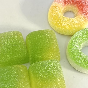 Op de afbeelding: Een assortiment kleurrijke gummy snoepjes. De afbeelding toont groene, vierkante gummies en ringvormige gummies in geel, rood en groen. De snoepjes zijn bedekt met een suikerachtige substantie, waardoor ze een berijpt uiterlijk hebben.