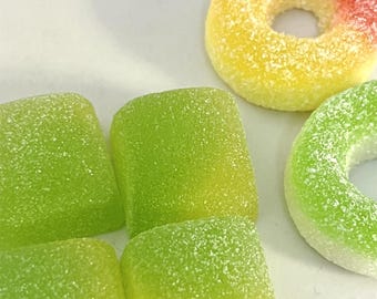 Op de afbeelding: Een assortiment kleurrijke gummy snoepjes. De afbeelding toont groene, vierkante gummies en ringvormige gummies in geel, rood en groen. De snoepjes zijn bedekt met een suikerachtige substantie, waardoor ze een berijpt uiterlijk hebben.