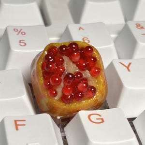 May include: A close-up of a small, red and yellow pomegranate replica. The replica is made of resin and has a realistic appearance. It is sitting on a white keyboard.