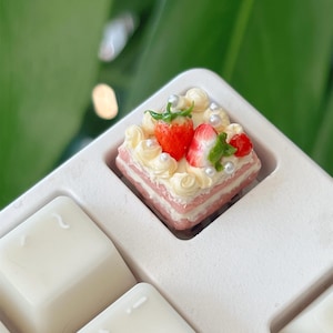 May include: A miniature strawberry cake with white frosting, a sliced strawberry, and pearl sprinkles sits in a white tray. The cake has pink layers and is surrounded by white wax cubes.
