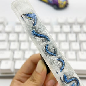 May include: A clear resin pen with blue and silver dragonflies embedded inside. The pen is held in a hand against a white keyboard.