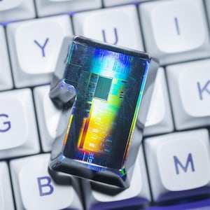 May include: A close-up of a computer keycap with a reflective, multi-coloured microchip on top. The keycap is grey and sits on a white keyboard with purple lettering. The microchip has a complex, colourful internal structure.