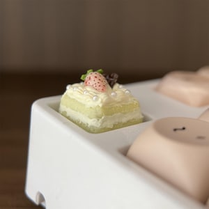 Può includere: Una tortina quadrata con una fragola sopra, adagiata in un vassoio bianco. La torta ha strati gialli e bianchi, decorata con glassa bianca e piccole perle. È presente anche un piccolo pezzo di cioccolato.