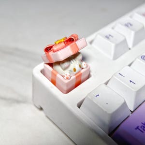 Puede incluir: Un tapón de tecla en forma de caja de regalo rosa y blanca con una figurita de gato blanco en su interior. El tapón de tecla está en un teclado blanco.
