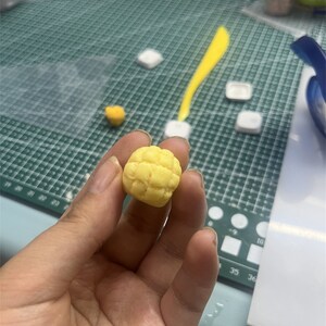 May include: A yellow, textured, round, handmade polymer clay food charm shaped like a pineapple bun.