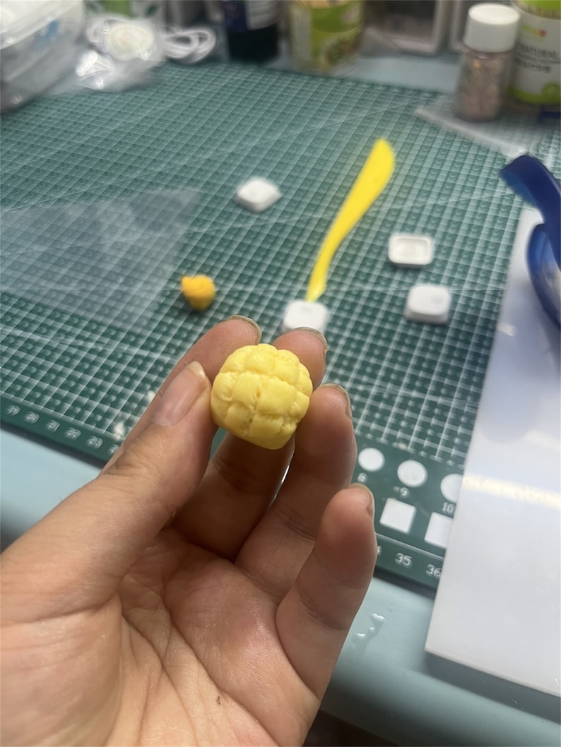May include: A yellow, textured, round, handmade polymer clay food charm shaped like a pineapple bun.