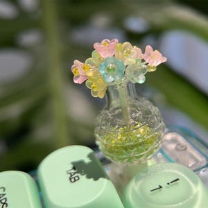 May include: A miniature glass vase filled with green glitter and topped with a bouquet of colorful, translucent flowers. The vase sits atop a keyboard with light green keys, including the "TAB" and "1" keys.