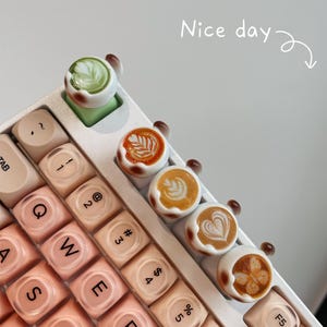 Puede incluir: Primer plano de un teclado mecánico con teclas personalizadas con temática de café. Las teclas están diseñadas para parecerse a arte latte, con diferentes diseños de café. El teclado tiene un marco blanco y teclas rosas. El texto "Nice day" está escrito en cursiva.