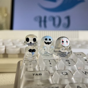 Könnte beinhalten: Drei durchsichtige Plastikfiguren mit verschiedenen Gesichtern auf einer weißen Tastatur. Die Figuren sitzen auf den Tasten der Tastatur. Die Tastatur hat durchsichtige Tasten mit schwarzer Beschriftung. Die Figuren sind ein Geist, ein Schädel und eine Figur mit einer roten Maske.