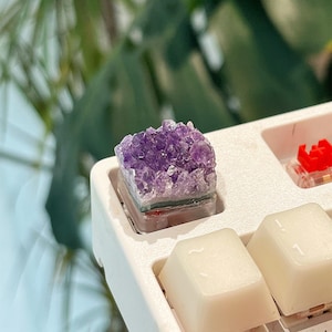 Puede incluir: Una tecla personalizada para teclado mecánico que presenta un racimo de cristales de amatista púrpura en bruto y dentados sobre una base cuadrada, colocada en un marco de teclado blanco junto a teclas color crema e interruptores rojos.