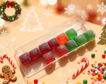 Op de afbeelding: Een doorzichtige plastic doos gevuld met kleurrijke, gesuikerde gummy snoepjes. De snoepjes zijn rood, oranje en groen, gerangschikt in rijen. Kerstversieringen, waaronder een krans, sneeuwvlokken en een peperkoeken man, omringen de doos.
