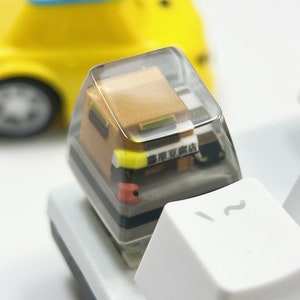 May include: A clear resin keycap with a miniature yellow car inside. The car has a blue roof and the text "旅館案内" is visible on the side of the car.
