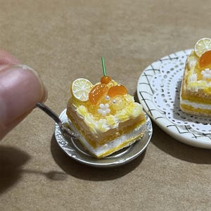 Puede incluir: Un pastel cuadrado en miniatura de cítricos sobre un pequeño plato plateado, cubierto con glaseado blanco, gajos de naranja, una rodaja de limón y un tallo verde diminuto. Una cuchara plateada descansa contra el pastel de capas amarillas y blancas.