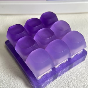May include: A set of nine purple translucent keycaps for a mechanical keyboard. The keycaps are arranged in a 3x3 grid and are sitting on a purple plastic base.