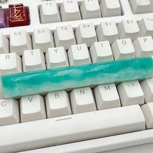 Puede incluir: Un teclado blanco con una tecla de resina de color verde azulado y blanco. La tecla es un reemplazo personalizado para una tecla de teclado estándar.