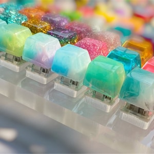 Può includere: Primo piano di una collezione di tasti traslucidi e colorati. I tasti sono disposti in file su un espositore in acrilico trasparente. I tasti sono realizzati in resina e presentano una varietà di colori e motivi, tra cui blu, verde, rosa e giallo.