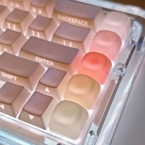 Könnte beinhalten: Eine mechanische Tastatur mit transparentem Rahmen und hintergrundbeleuchteten Tasten in sanften Rosa-, Pfirsich- und Cremetönen. Die Tasten tragen weiße Beschriftungen wie BACKSPACE, ENTER und SHIFT und leuchten von unten.
