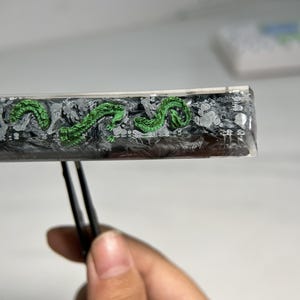 May include: A close-up of a black and white resin rectangle with a green dragon design. The rectangle is being held by a pair of tweezers.