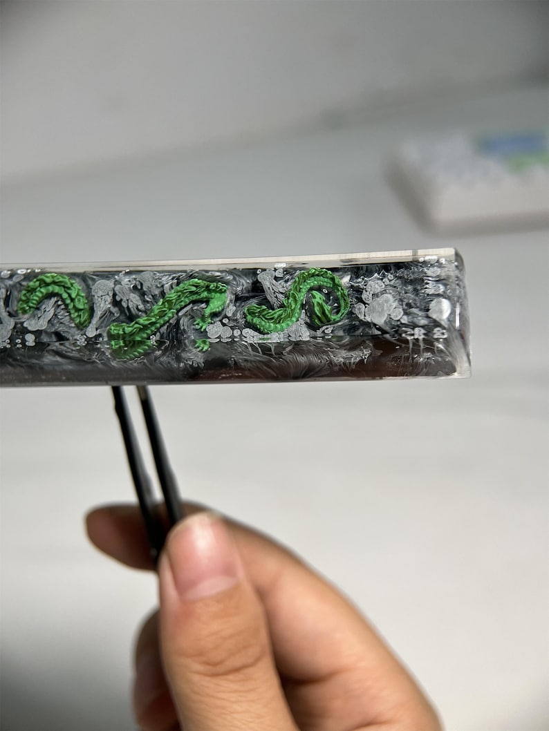 May include: A close-up of a black and white resin rectangle with a green dragon design. The rectangle is being held by a pair of tweezers.