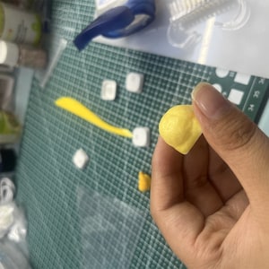 May include: A small, yellow, handmade clay bead. The bead is round and has a slightly uneven surface. It is being held in a person's hand.