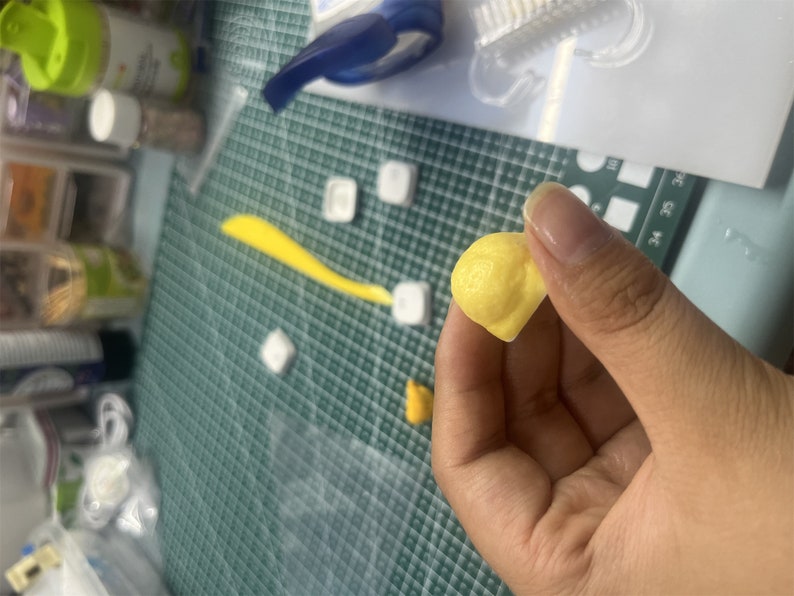 May include: A small, yellow, handmade clay bead. The bead is round and has a slightly uneven surface. It is being held in a person's hand.
