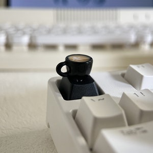 Op de afbeelding: Een miniatuur zwart koffiekopje met een bruine vloeistof en een wit ontwerp zit bovenop een zwarte toetsdop op een wit toetsenbord.