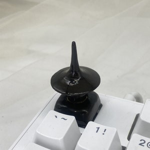 May include: A glossy black decorative object with a conical top and a wide, flat base. The object is placed on a white keyboard, with the keys "~", "1", and "2" visible. The background is a soft, white fabric.