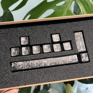 May include: A set of custom keyboard keycaps with a carbon fiber pattern. The keycaps are arranged in a wooden box with a black foam insert. The keys include the letters A, S, D, W, and an Enter key.