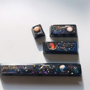 May include: A set of black resin keycaps with a space theme. Each keycap displays a miniature galaxy design, including planets, stars, and cosmic dust. The keycaps are in various sizes and shapes, with rectangular and square designs.