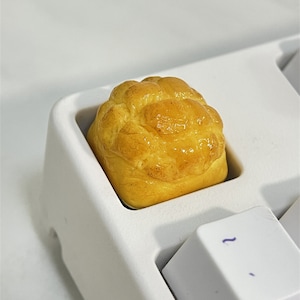 May include: A close-up of a yellow and brown bread-shaped keycap on a white keyboard.