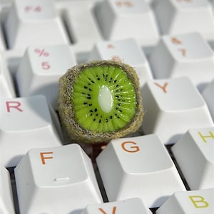 Puede incluir: Una rodaja de kiwi verde con piel marrón peluda se coloca sobre un teclado blanco. El kiwi está centrado en las teclas F y G.