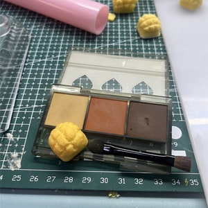 May include: A clear plastic eyeshadow palette with three different shades of brown eyeshadow. The palette is open and there is a small yellow sponge applicator brush next to it. There are also several small yellow pastries on a white surface.