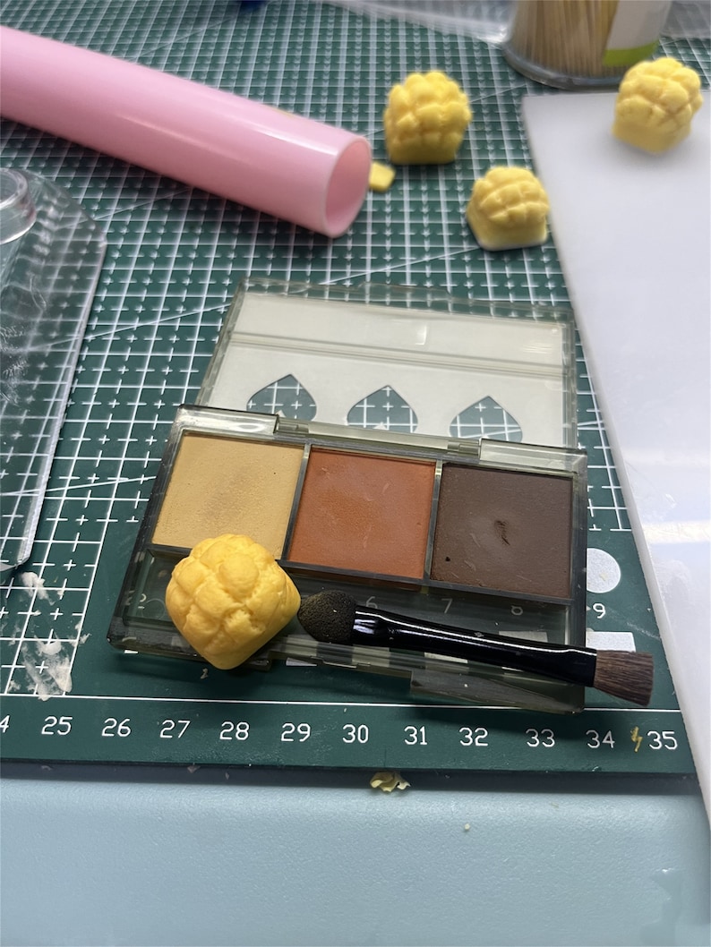 May include: A clear plastic eyeshadow palette with three different shades of brown eyeshadow. The palette is open and there is a small yellow sponge applicator brush next to it. There are also several small yellow pastries on a white surface.