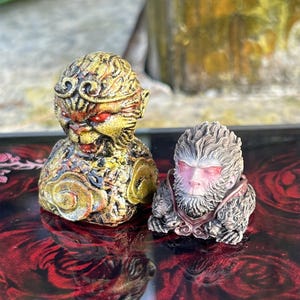 Może przedstawiać: Dwie małe, szczegółowe figurki małp. Małpa po lewej stronie jest złota z czerwonymi oczami i koroną. Małpa po prawej stronie jest szara z czerwonymi oczami i brodą. Obie małpy siedzą na czerwono-czarnej powierzchni.