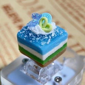 May include: A miniature cake with blue, white, green, and brown layers, topped with white frosting, a green swirl, a blue bear, and white sprinkles. The cake is on a clear plastic base.