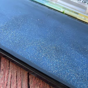 K&ouml;nnte beinhalten: Nahaufnahme einer dunkelblauen Oberfl&auml;che mit einer feinen Schicht Glitzer. Die Oberfl&auml;che ist glatt und reflektierend, mit einem leichten Glanz.